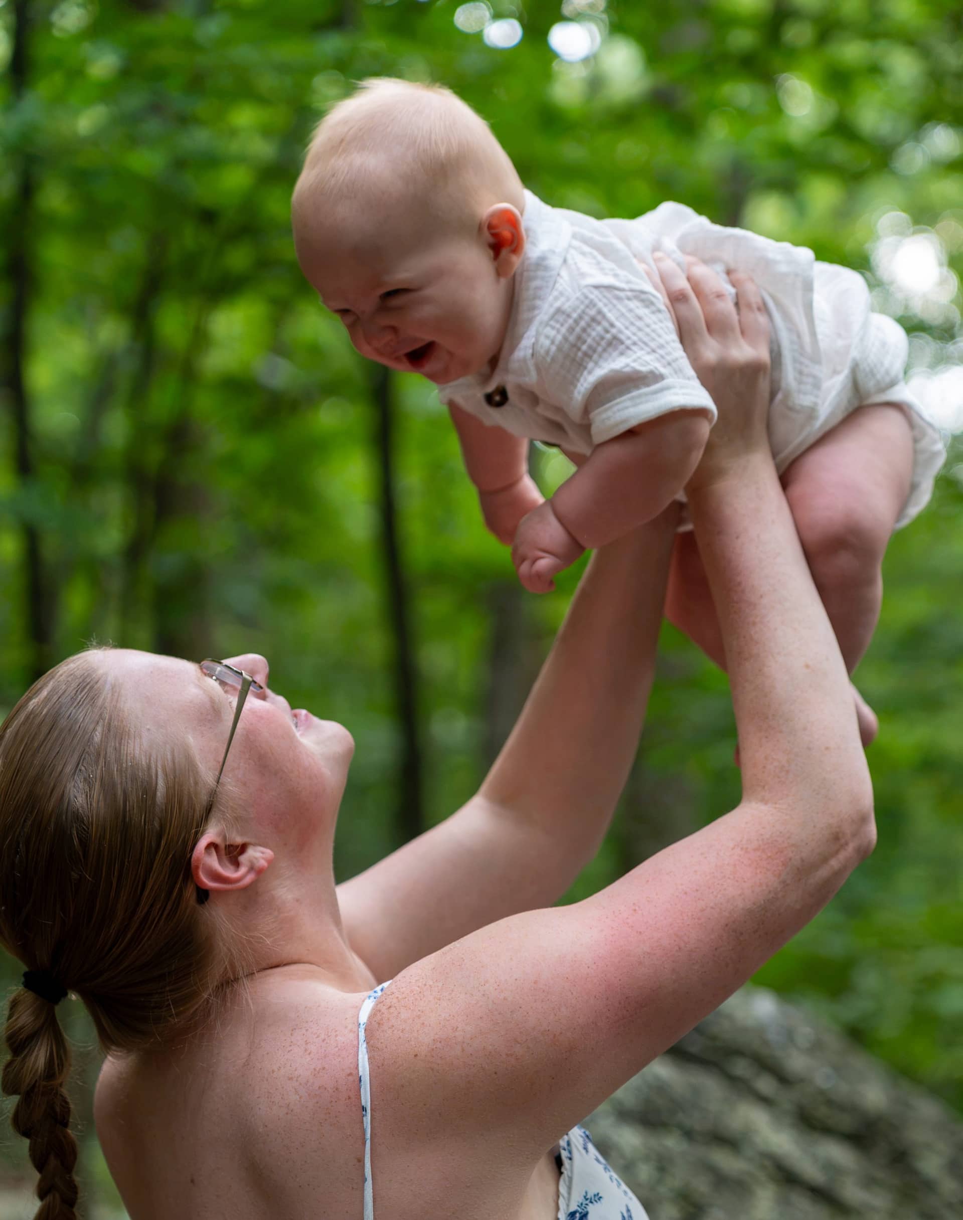 A mother lifting her laughing baby above her head in dappled woodland light