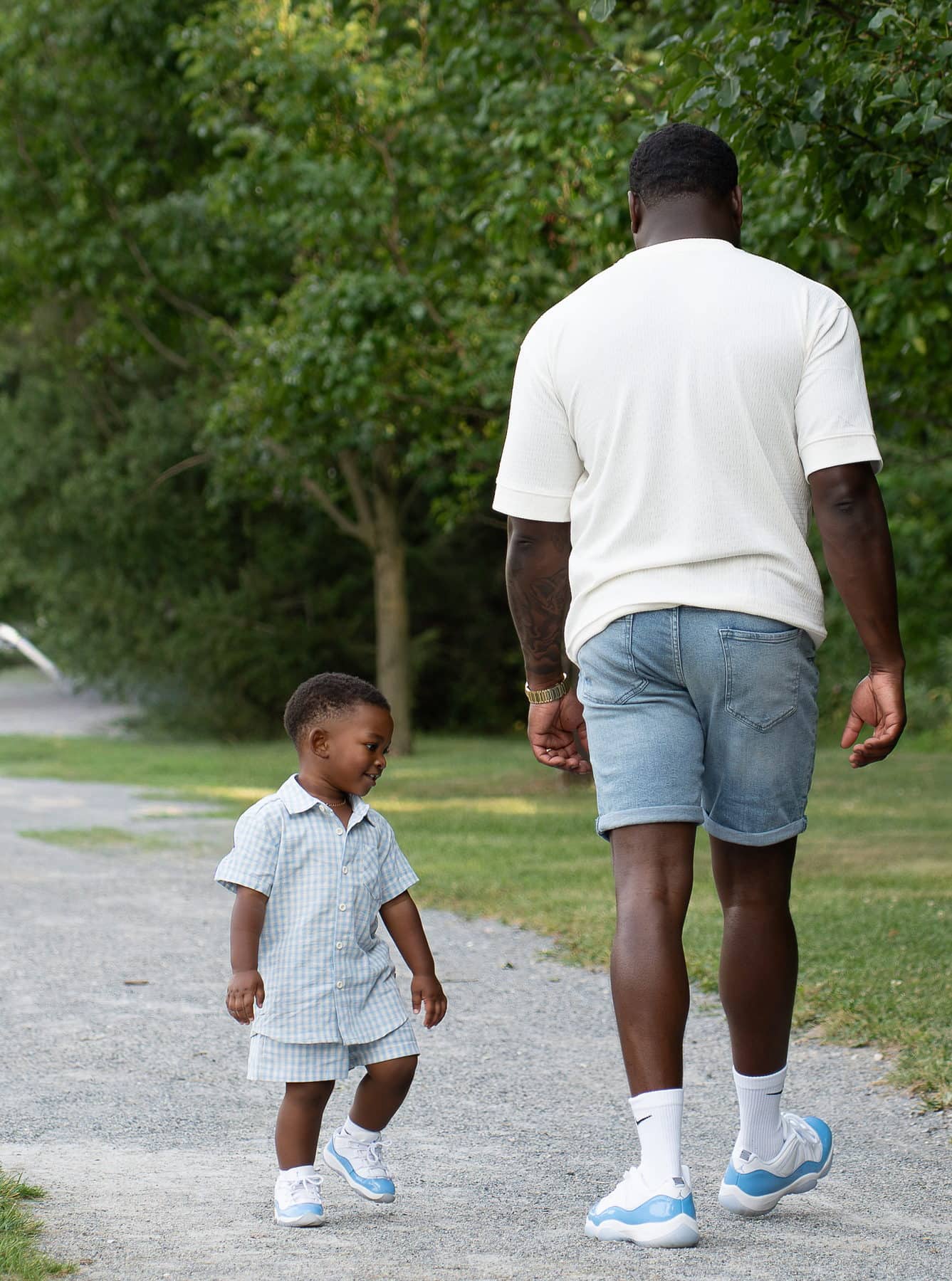 A toddler walking a park path beside his father, both seen from behind