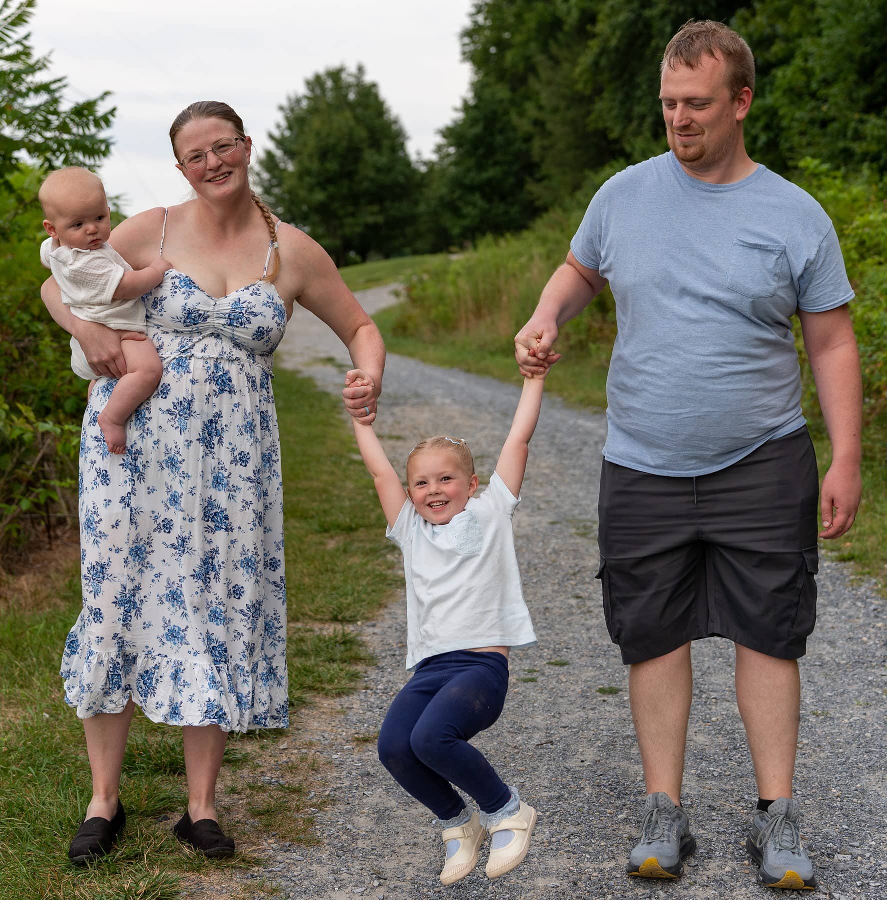 A mother and father swinging their daughter between them on a gravel path, baby on mum's hip