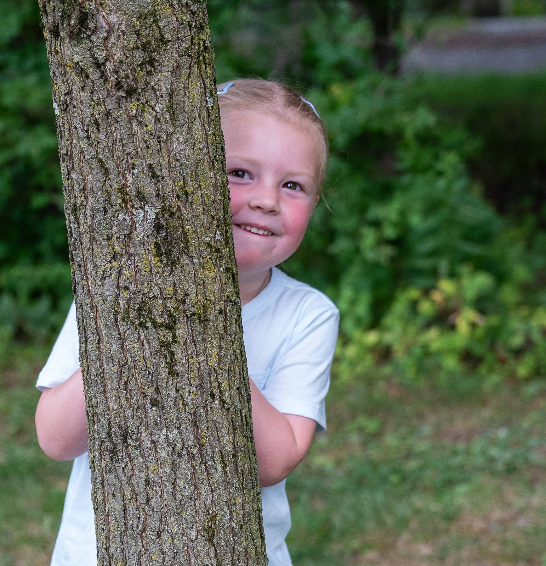 A young girl half hidden behind a tree trunk, peeking out