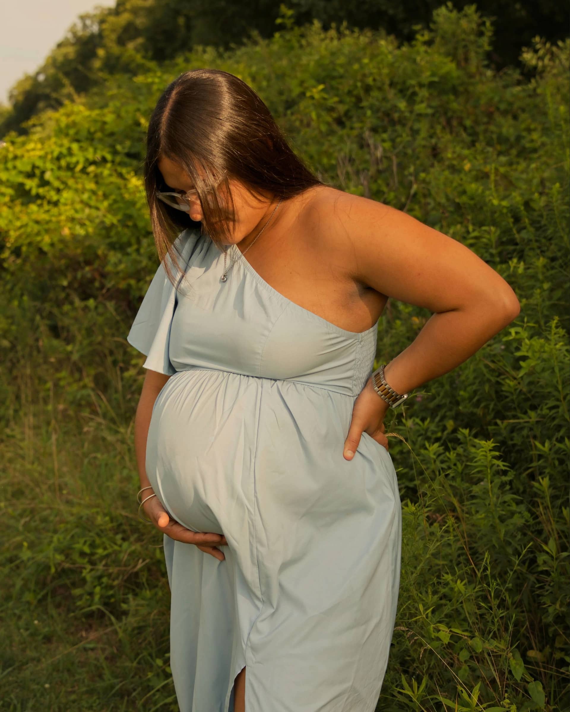 An expectant mother in a sage green dress, hands resting on her bump, surrounded by summer greenery