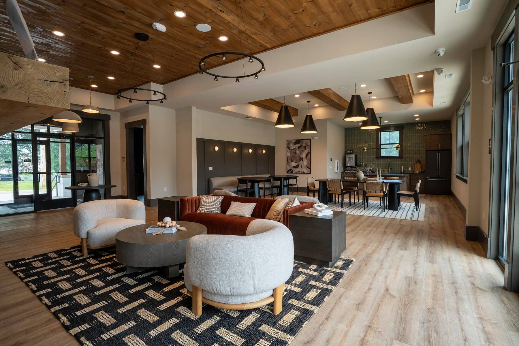 A clubhouse lounge with a rust velvet sofa and boucle chairs on a graphic rug, opening through to the community kitchen