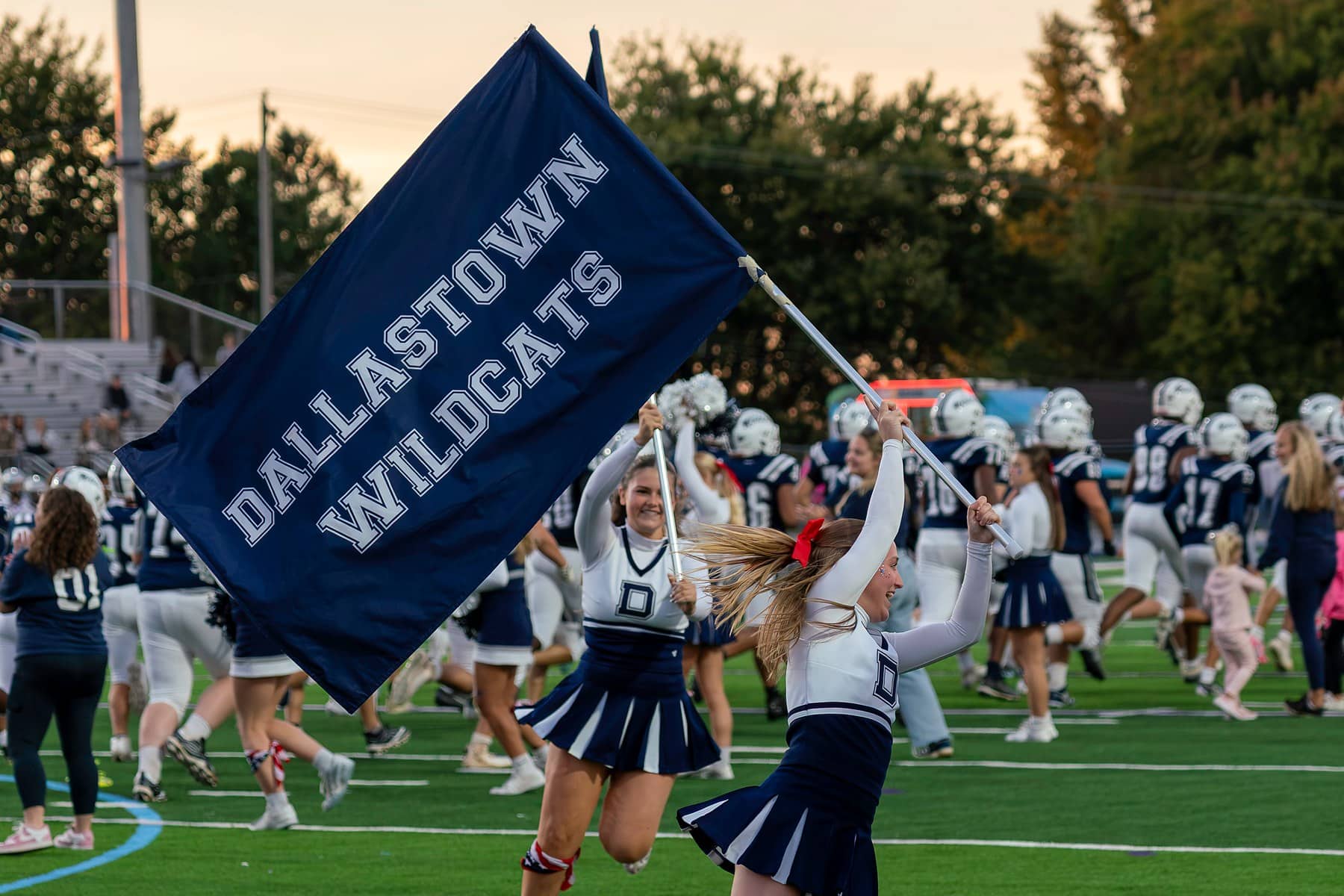 A cheerleader carrying the Dallastown Wildcats flag across the field at dusk as the football team runs out behind her