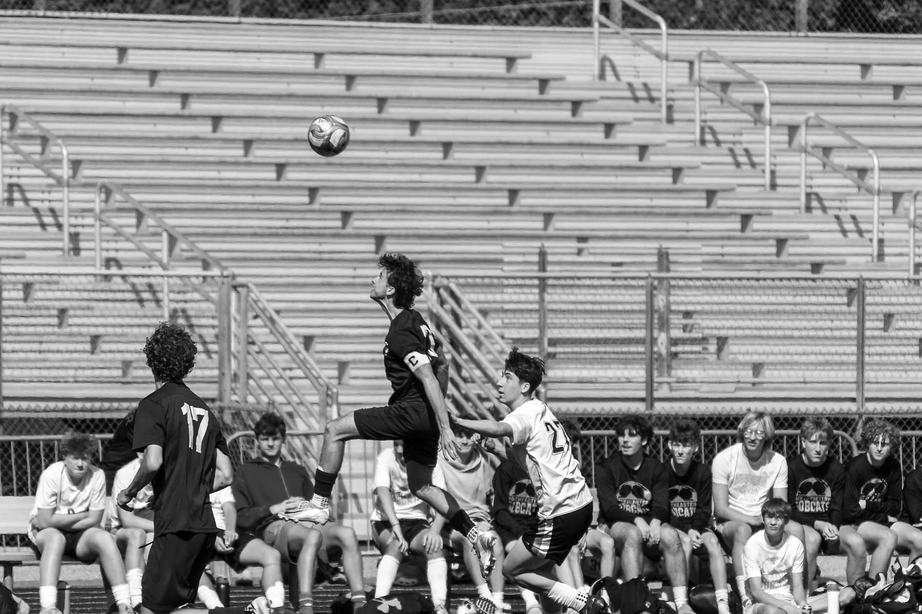 Two high school soccer players rising together for a header while the opposing bench watches, in black and white