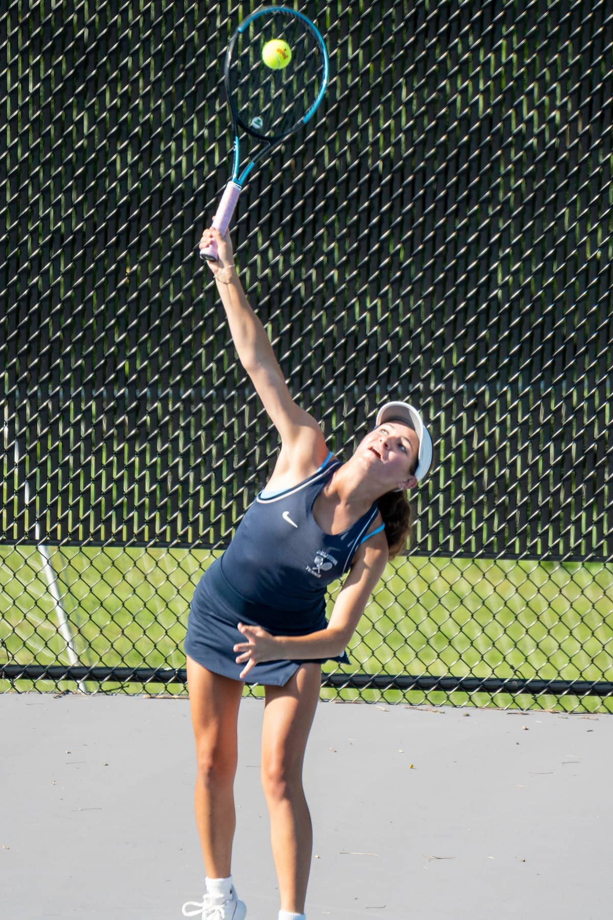 A high school tennis player at full extension on a serve, the ball framed inside the racket