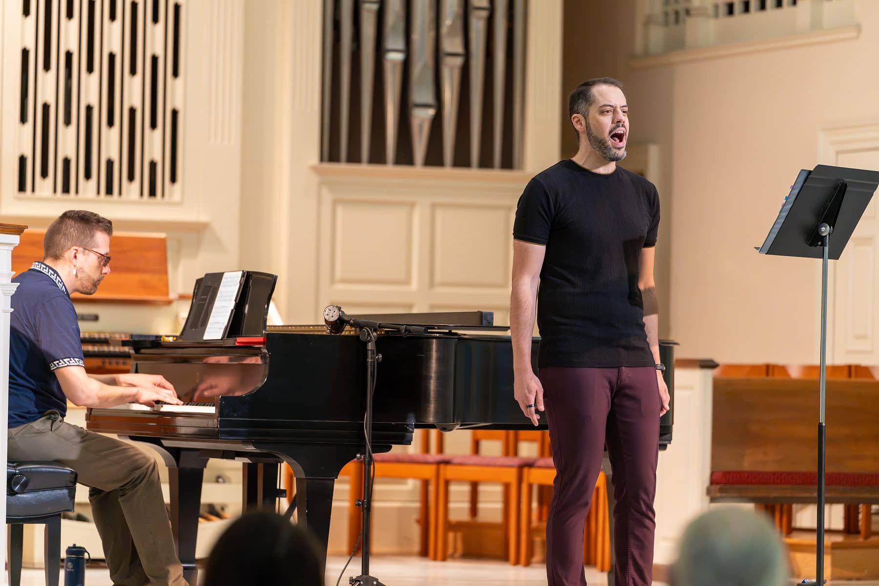 A tenor singing mid-phrase beside his accompanist at a grand piano, organ pipes behind them